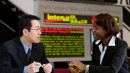 An Asian and Black student in suits hold pens while talking in front of an electronic stock ticker sign.