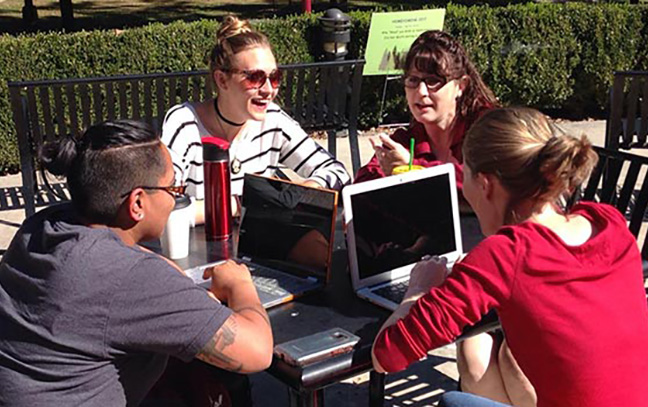 English graduate students gather around a table outdoors. There are laptops on the table.