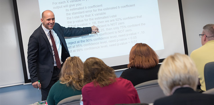 A professor stands in front of a projector screen, pointing something out to the MBA class