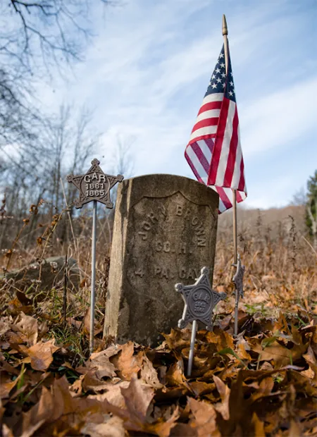 An old gravestone with military markers and an American flag sticking in the ground next to it, surrounded by fall leaves