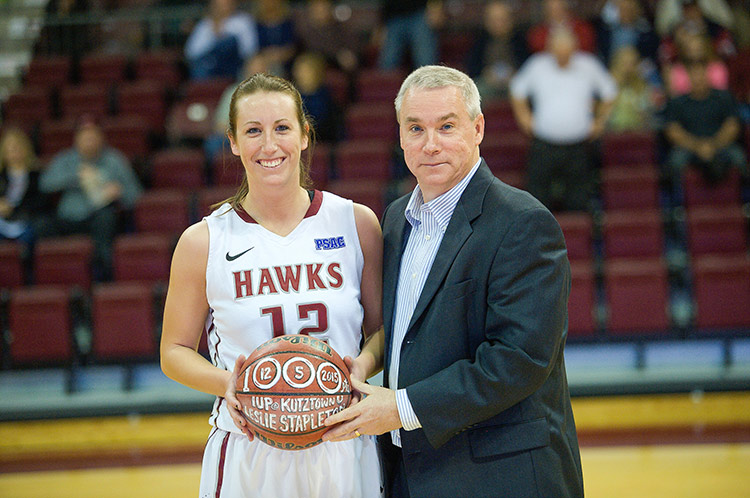 Leslie Stapleton, in a white basketball uniform, stands smiling on the basketball court next to her coach, Tom McConnell, who is in a dark suit and light blue dress shirt. She is holding the ball from her 1,000th point. It has her name on it. The crowd is seen blurred in the stands in the background.