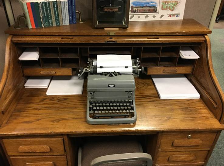 A wooden roll-top desk with books, paper, and an old typewriter resting on it