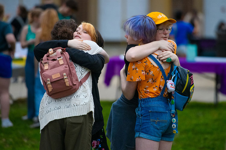 Two sets of female students hug one another in the Oak Grove, with other people visible in the background, along with event display tables.