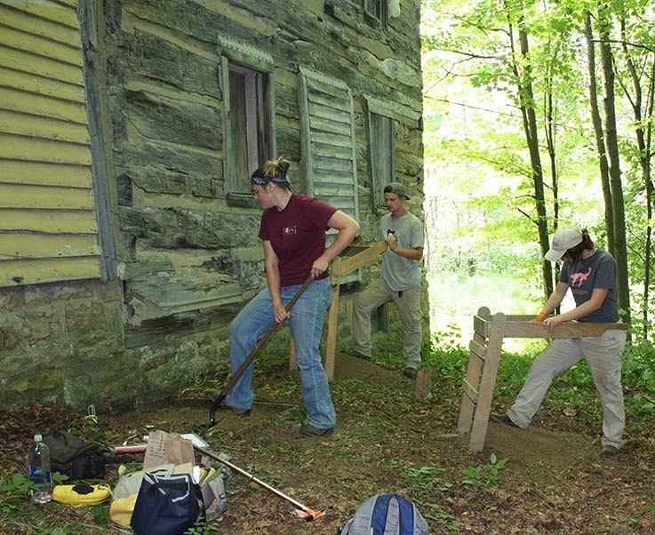 An archeology student digs at the base of an old historic building near the woods, while two other students operate rocker screens to separate finer materials from larger ones.