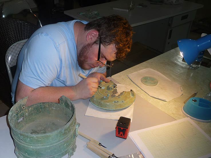 An archeology student sits at a table with a desk lamp illuminating as they carefully work on an archeological artifact.