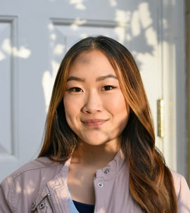 a young woman standing in front of a white door faces the camera as the shadows of tree branches overlap her