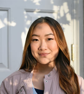 a young woman standing in front of a white door faces the camera as the shadows of tree branches overlap her