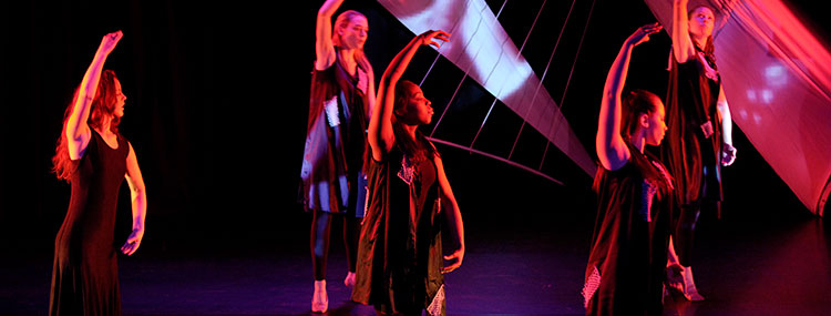 Five dancers dressed in black dance with one arm curved above their heads on a darkened stage lit with red light