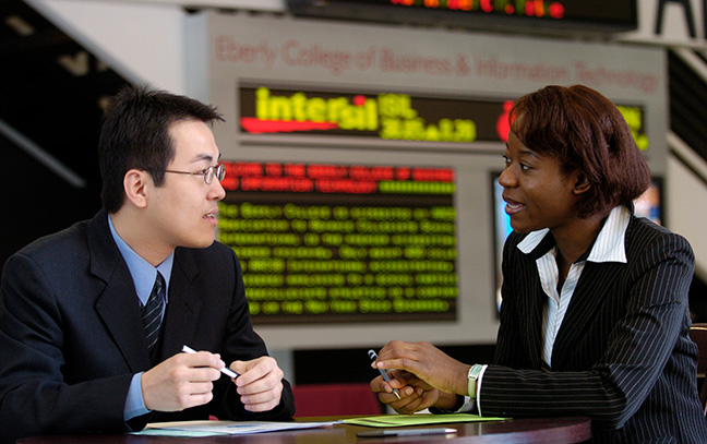 Two graduate business students talking in front of a stock ticker