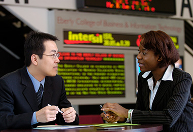 Two graduate business students talk in front of a stock ticker