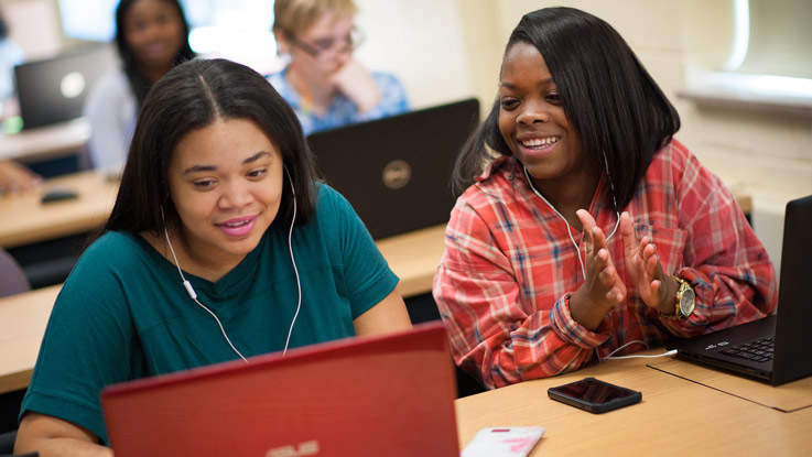 Two female students work together on a laptop in a classroom