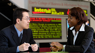 An Asian and Black student in suits hold pens while talking in front of an electronic stock ticker sign.
