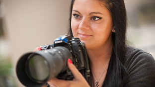 A female photography student smiles and holds a professional long-lens camera.