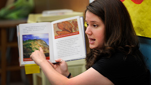 A student holds up a book and points to its illustration while discussing it. 