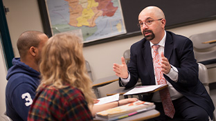 A professor wearing a suit and tie explains concepts with two students in a classroom as a map of France hangs on the wall.