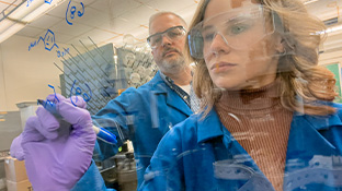 Professor Fair and student wearing gloves and safety goggles draw molecular diagrams on a pane of glass in a lab.