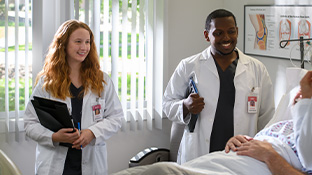 Smiling nursing students wearing lab coats listen to a patient who lays in a hospital bed.