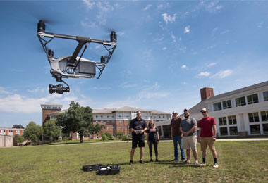 A remote-controlled geography drone flies above a field on campus has a group of students with their professor look on. 