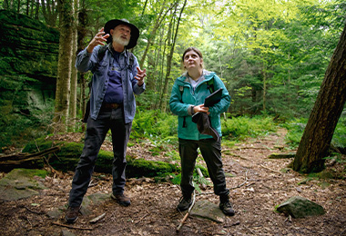 A geoscience professor gestures and looks up while discussing with a student along a scenic trail in Bilge's Rocks.