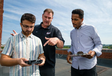 Professor Benhart points at a flying drone a student holds while conducting a parking analysis study. Student Trajan Jones looks on.