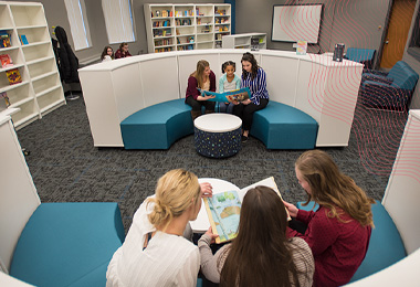 Female students reading books to children while sitting in a modern circular seating area.
