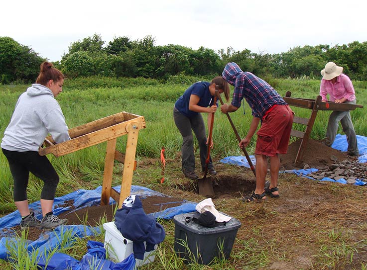 Two archeology students use shovels while digging into a hole, while two other students operate rocker screens to separate finer materials from larger ones.