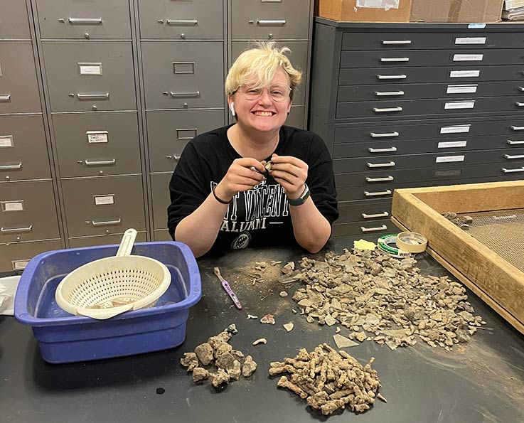 An archeology student sits at a table in a lab/storage room, smiling while sorting through material from a rocker screen.
