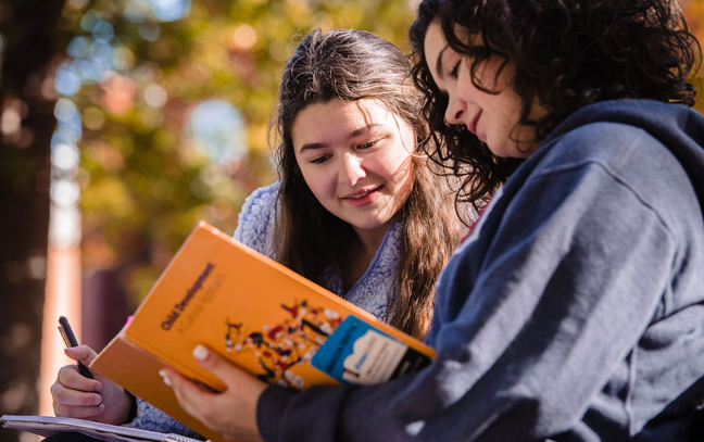 Two female students look at a book in the Oak Grove
