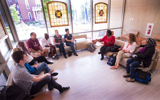 a large group of students and a professor are seated in a circle having a discussion in class