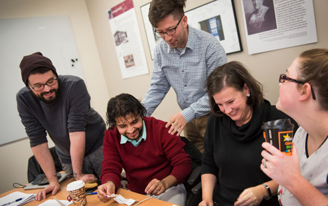 a small group of students gathered around a table with a professor talking and smiling