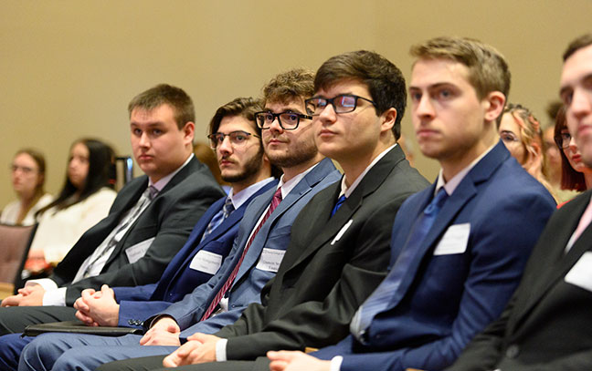 a line of students seated listening to a speaker at business day