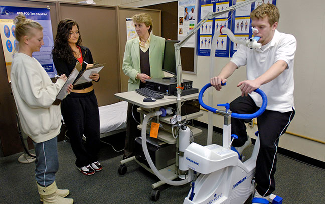 a student rides a stationary bike while others monitors, with a professor watching at the back