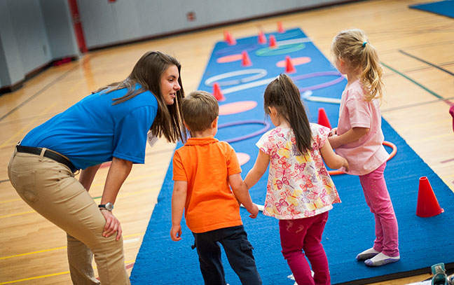 a female student teacher leans down and talks to three small children in a gym