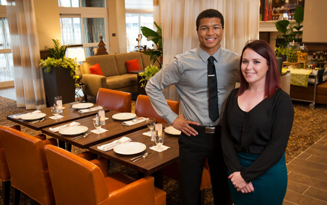 Three students stand side by side, smiling at the camera in a hotel setting