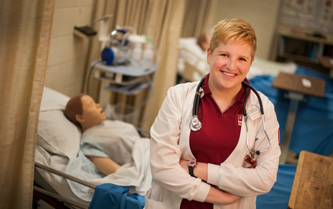 a nursing student standing in a simulation lab smiling at the camera