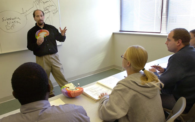 a teacher stand at the front of a small classroom talking to students