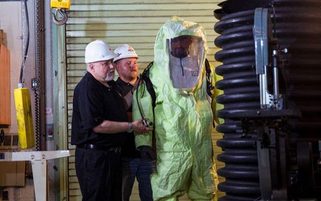 two men help another person into a hazmat suit in an industrial setting