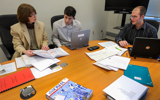 a group of three people seated at a table with laptops look over documents