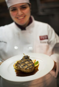 female culinary student holds out a plate with a grilled pork chop on it