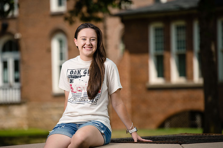 Chrissy smiles sitting outside Sutton Hall on a sunny day. 