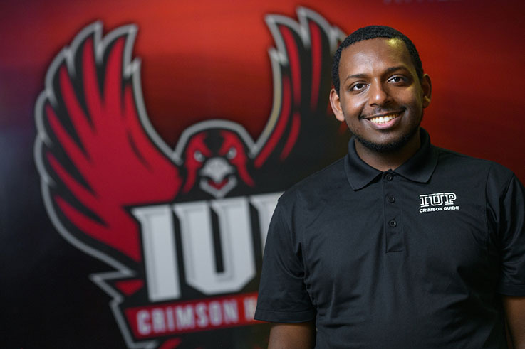 Enoch smiles in the KCAC in front of the athletic Crimson Hawk logo.