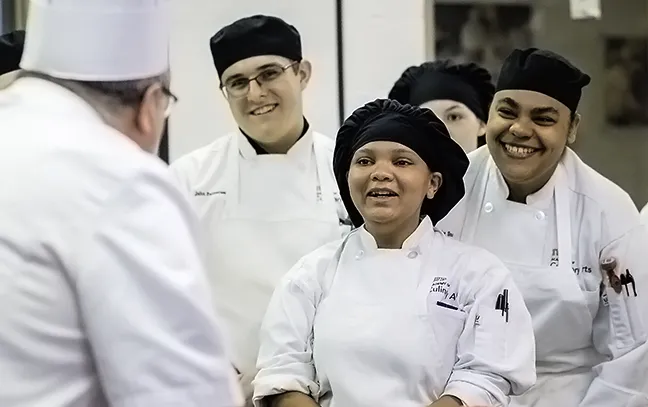 culinary students facing their instructor and smiling while in a kitchen