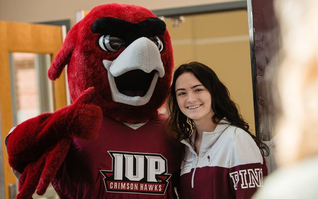 a student posing with the IUP crimson hawk mascot as he gives a thumbs up