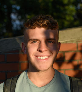 a young man standing in front of a brick wall faces the camera as the shadows of tree branches overlap him