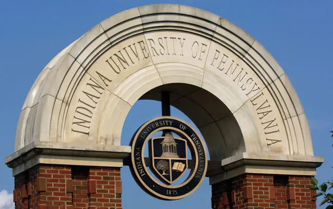 a bright blue sky with a brick and concrete arch with a metal IUP academic seal mounted under the top