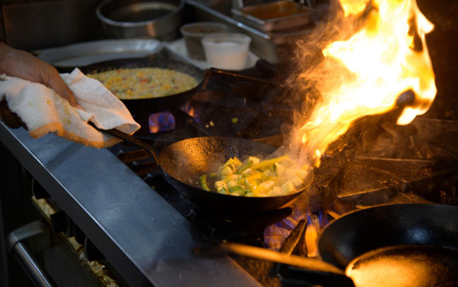 a hand using a cloth to flip flaming food in a hot frying pan over a gas stove