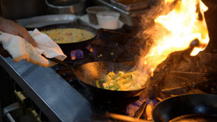 a chef uses a cloth to flip flaming food in a hot frying pan over a stove