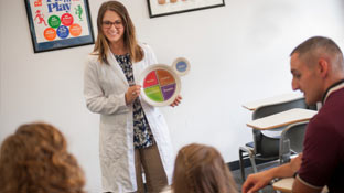 a teacher at the front of a room holding up a food chart to a group of three people