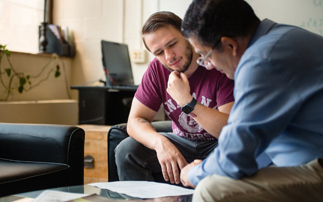 a professor and student sitting next to each other going over papers on a coffee table.