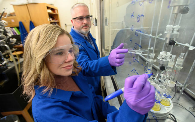 a young woman in a blue coat in a lab writes on a clear acrylic panel while a man standing next to her instructs her.